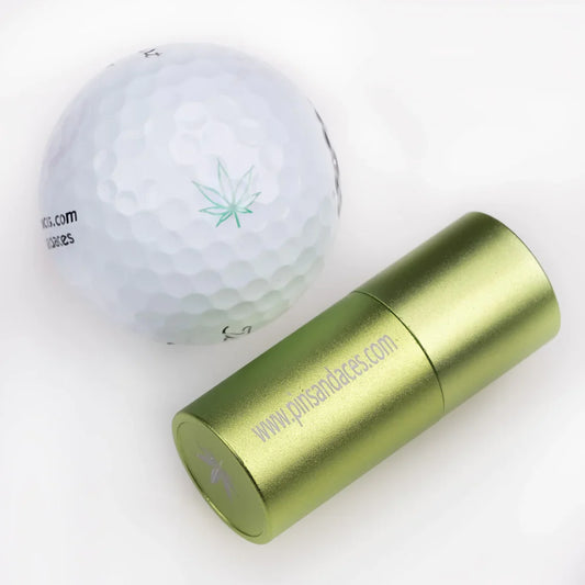 Alt-text: "En vit golfboll med en grön cannabisblad-logotyp bredvid en metallisk guldgrön golfbollstämpel med webbadressen på, båda på en vit bakgrund.