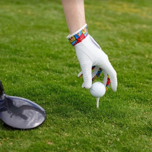 En hand iförd en vit golfhandske som håller i en golfboll. Handsken har detaljer med färgglada mönster på remmen runt handleden. I bakgrunden syns grön gräs. Till vänster i bilden skymtar en golfklubba.