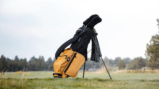 En gul och svart golfbag står på ett stativ på en dimmig golfbana, omgiven av gräs täckt av dagg