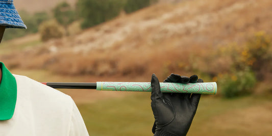 Golfspelare i förgrunden håller en golfklubba med en detaljerad grönt mönstrad golfgrepp, iklädd en svart golfhandske och en blå och grön bukethat, med en suddig golfbana i bakgrunden.