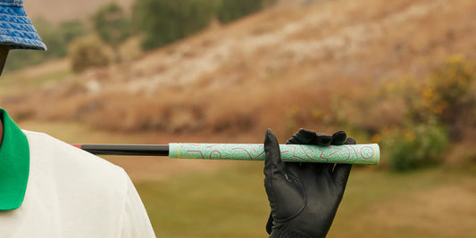 Golfspelare i förgrunden håller en golfklubba med en detaljerad grönt mönstrad golfgrepp, iklädd en svart golfhandske och en blå och grön bukethat, med en suddig golfbana i bakgrunden.