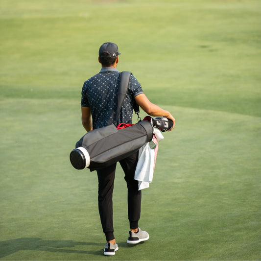 Bakifrånvy av en golfspelare iklädd en mörk golfkeps, en mönstrad skjorta och byxor, som bär en grå golfbag över axeln medan han går över en grönskimrande golfbana.