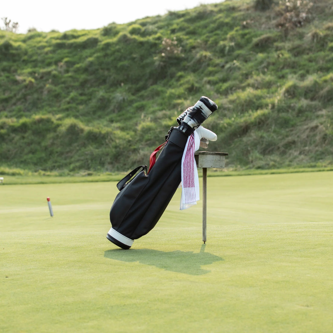 En svart och röd golfbag står på en grön golfbana med en vit golfhandduk som hänger från en av klubborna, nära ett golfhål med flaggstång i bakgrunden och en kulle med frodig vegetation.