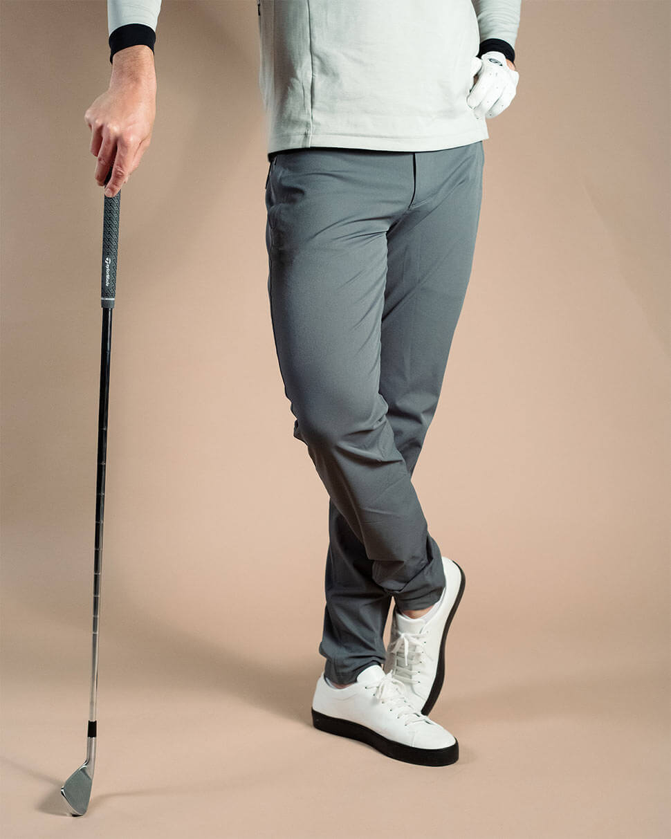 En person iklädd grå golfbyxor, vita golfskor och en vit golfhandske håller i en golfklubba, avbildad från midjan ner på en neutral beige bakgrund