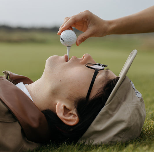 närbild på ett ansikte som ligger ner på en golfbana och har en pegg placerad i munnen. En hand lägger en golfboll på peggen.