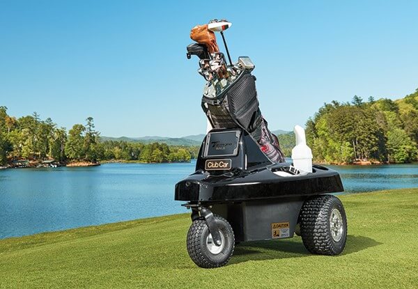 En fjärrstyrd golfvagn av märket Club Car, lastad med en golfbag full av klubbor, står på en frodig golfbana med en stilla sjö och skog i bakgrunden.