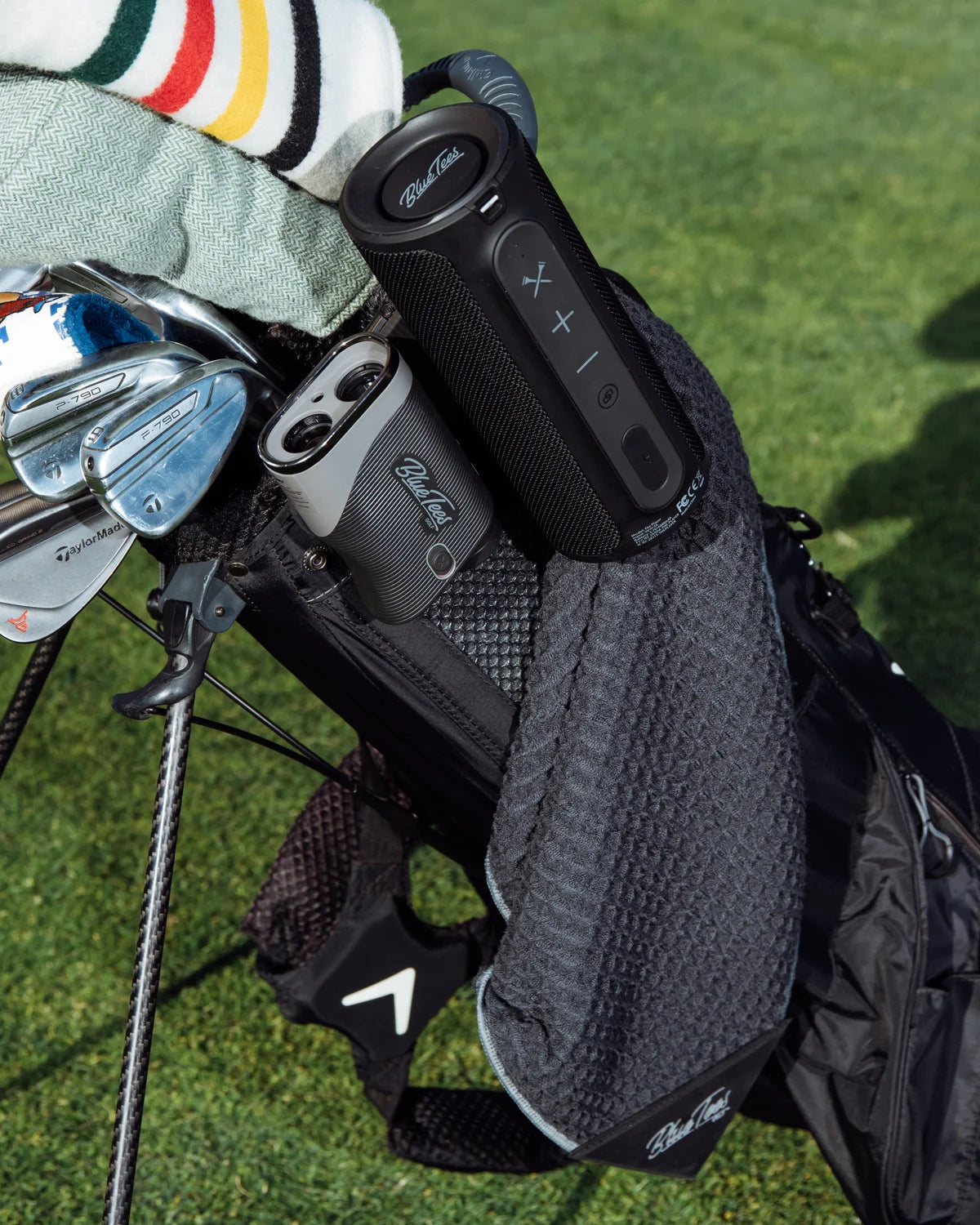 En svart golfbag, stående på en golfbana, svart golfhanduk hängande på golfbagen, på bagen finns även en golfkikare